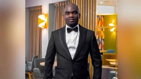 @Blesspixels Legton Zulu standing in a black suit and bow-tie with a white shirt. He is looking directly at the camera and smiling.