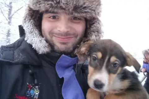 Bernard Mongan posing for a photo with a dog. Mr Mongan is wearing a fluffy hat.