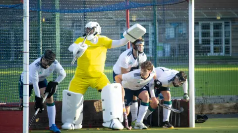 Chichester Hockey Club The men's first team waiting in their goal for a corner, the keeper is wearing a yellow kit with pads and there's four players in the goal mouth ready to rush out wearing white kits and protective masks