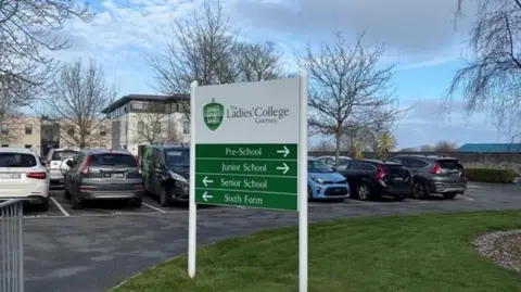 BBC A white and green sign held up with two pillars reads THE LADIES' COLLEGE GUERNSEY. Behind it is a car park which looks full and behind that is the college which appears to be three to four storeys and modern in design. 