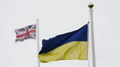 PA Media A Ukrainian flag flying on a pole against a grey sky. In the background there is a Union Jack flag flying out of focus.