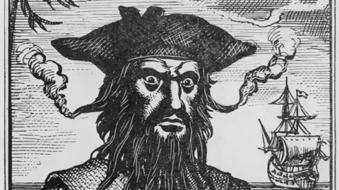 A woodcut engraving of Blackbeard, showing a man with a long black beard, and smoke coming off some of the hairs. A sailing ship is visible in the background.