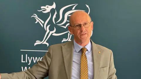 Llyr Gruffydd speaking during a press conference. He is wearing a beige blazer, a light blue shirt and yellow tie. He is bald and wears round glasses.