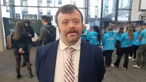 James Martin has short brown hair and a moustache and is wearing a navy suit jacket, white shirt and striped tie. He is standing in front of a number of students  