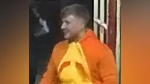 A young man with light hair in an orange costume, in a grainy CCTV image.