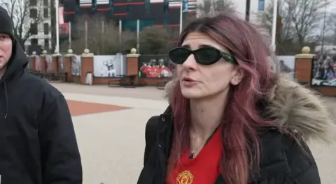 Woman with pink hair and united t shirt. She wears dark sunglasses and a black, fur-hooded parka.
