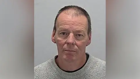 A mugshot of a man with short black hair wearing a grey jumper with a black top underneath.