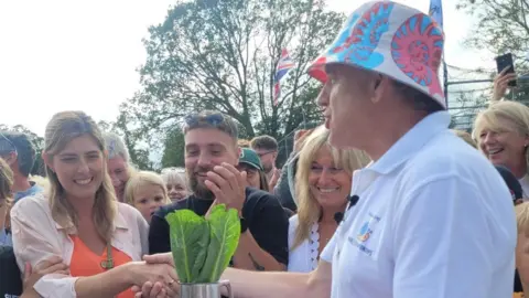 Stephen Huntley/BBC A man in a white T-shirt and a hat, shaking a woman's hand, holding a tankard with lettuce in. Other people are standing around. 