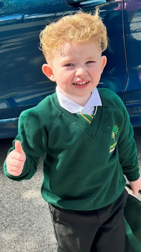 A boy wearing a green jumper with his thumb up and smiling towards the camera