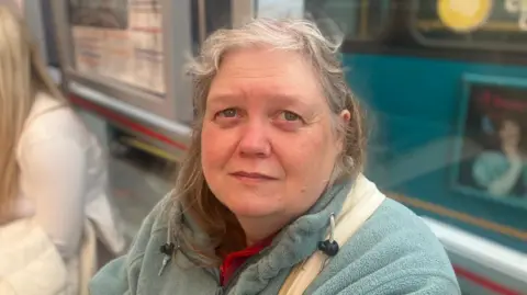 Elizabeth Baines/BBC A woman with grey hair sits at a bus stop, wearing a blue fleece with a white bag over her shoulder. Behind her, a teal bus is blurred.