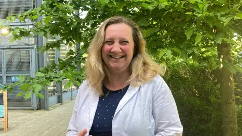 Lucy Nethsingha wearing a white blouse with a blue and white dress. She has shoulder length blonde hair and is smiling at the camera. 