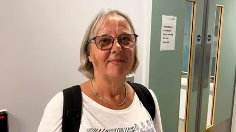 Renate Kennedy in A&E in Bournemouth. She has shoulder length blonde hair and is wearing a white top with the black straps of a bag over her shoulders. She is wearing glasses and a silver necklace. She is standing in front of a white wall with green double doors.