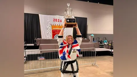 Becky Sheppard Becky Sheppard is wearing a martial arts uniform, which consists of a white T-jacket, trousers and black belt. She is holding a silver trophy, which has a black plaque on the front of it, above her head.