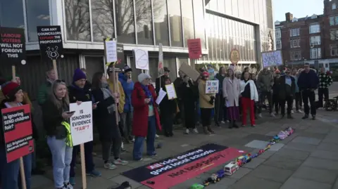 A large group of protesting parents, carrying placards, stand outside Hove Town Hall. A chain of toy cars and other vehicles has been placed in front of them.