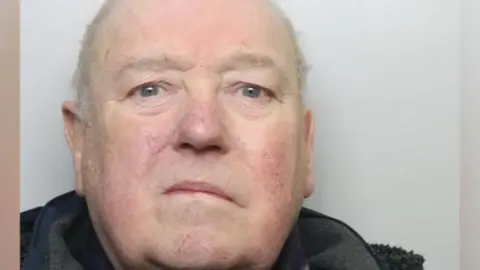 A police mugshot of a man in his eighties, mainly bald but with wispy white hair. He is wearing a navy coat and staring directly into the camera.