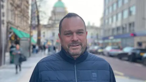 A man in a navy winter jacket looks at the camera. He has short dark hair and a dark short beard. Behind him is Belfast city hall and the high street. 