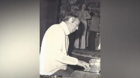 Daniel Hicks A black and white image of a man playing the piano. He wears a turtleneck jumper and glasses. 