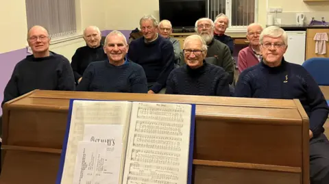 Group of 10 of the 14 men who make up Filey Fisherman's Choir which meets at Filey Methodist Church every Saturday evening. The men in the picture are mostly wearing navy fisherman's ganseys. There is a piano in front with music for various sea-related hymns.