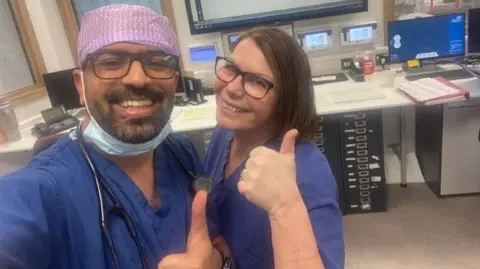 Two medical professionals, a man and a woman, in blue scrubs giving the thumbs up to the camera. The selfie is taken in an office environment, with desks and computers in the background. The man wears glasses and has a beard. His hair is covered by a pink bandana-style head covering. The woman has dark brown hair in a bob and is also wearing glasses.