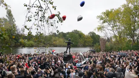 PA Media A large crowd gather round a statue, releasing love heart-shaped balloons