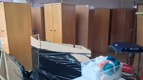 Help Ukraine BAMK UK Community Interest Company (CIC) A large amount of items in a room, including wardrobes, boxes in bin bags, and a box of wool. They wardrobes are all lined up. 