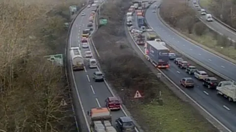 An ariel view from traffic camera of traffic on both sides of the A14, which is dual carriageway. There are wintry trees on the left and bushes on the central reservation.