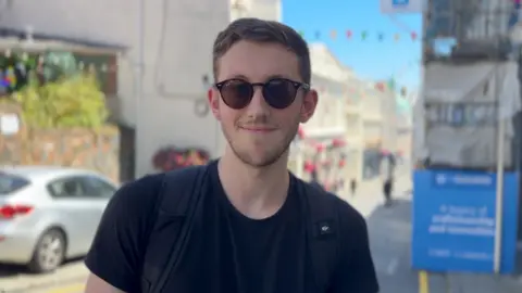 A man with sunglasses on smiling at the camera. He is wearing a black t-shirt and you can see the black straps of his ruck sack on his shoulders.