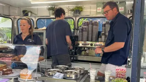Alex Pope/BBC Three people serving food from a mobile food van. A woman is by a glass case, a man has his back to the camera and another man is looking down. They are all wearing blue T-shirts. There is a large coffee machine behind them.