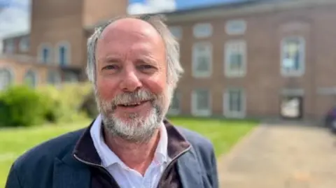 BBC Julian Brazil pictured bin front of county hall, wearing a navy blue jacket and white shirt. He has a grey and white beard with short receding hair.