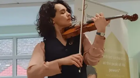 Woman with dark curly hair playing a violin, she has her eyes closed and wears a dark waistcoat and dusky pink shirt.