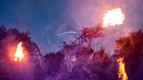 Paka the Uncredible A large dragon constructed out of large metallic beams and pipes is pictured at night. Fire is bursting out of its muzzle, with lit flames also at the tip of each of its wings. It is night time, and the tops of trees can be seen in the background. 