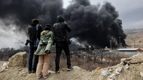 WANA/via Reuters People take photographs of smoke rising from burning fuel tanks in Tehran, Iran (8 March 2026)