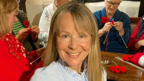 Tina Hill smiling, wearing a blue and white top. She has long hair and other women are behind her knitting by a table that has poppies on.