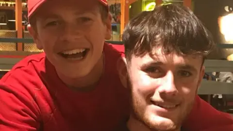 Abrahams Family A boy in a red jumper and a red cap smiles at the camera next to another boy with long brown hair and a light brown beard also smiling.