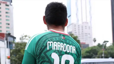 Photo of Maradona's green football shirt. The number on the back of the shirt is green. A man is wearing it with his back to the camera. He is standing in front of city centre traffic. 