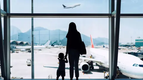 Getty Images A woman and child look out at an airport runway. They are silhouetted in shadow. In the distance there are a number of airplanes. 