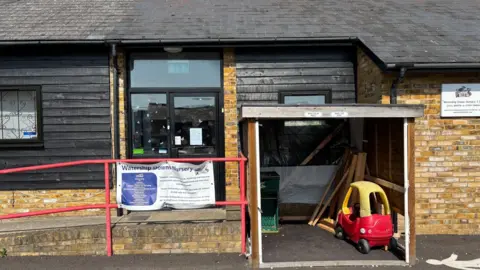 Jodie Halford/BBC A building with tan bricks and black cladding has signage on the outside saying Watership Downs nursery. A child's red and yellow Cozy Coupe car is in a shed in front of the building.