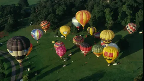 Richard Cardy Various hot air balloons coloured yellow, green, red, pink, white and blue in different combinations flying as seen from a balloon at an even greater height