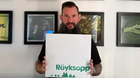 A man with dark hair and a long beard is holding a white vinyl with the word 'Röyksopp' in a green, stencilled format and green pixelated treetops. Behind him is various framed vinyls. 