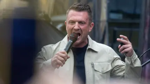 EPA Tommy Robinson, who has short brown hair and wears a collared beige jacket with a black t-shirt, is seen speaking into a microphone at the Unite the Kingdom rally