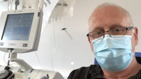 Andrew Bruce Andrew Bruce is wearing a light-blue face mask and looking at the camera. He has black glasses and is wearing a blue shirt with dots. He is hooked up to a bag filled with liquid and there is a grey monitor on the left. 