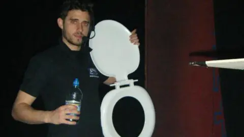 Ben Parsley Ben Parsley, holding a water bottle, wearing all black, with a headset on and holding a white toilet seat, he is backstage at a theatre. 