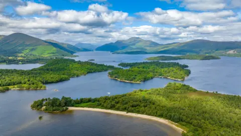 Alamy An aerial image of Loch Lomond, encompassing several of the lochs islands. The islands are beautiful shades of green and the water a rich blue. They lie in front of a rolling mountain range.