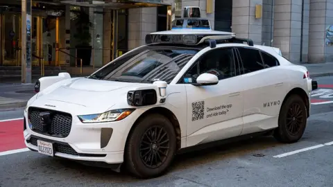 A white Waymo self-driving car with the words Waymo on the side, parked outside a building.