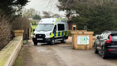 The picture shows a police van which has been parked across an entrance. There is a wooden block with a map on it to the right-hand side of the van. Behind it is a wooden log cabin.