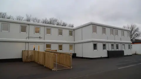 An exterior view of light-grey, two-storey "modular units", similar to pre-fab classrooms. A walkway, lined with wooden fencing attached to a metal handrail, leads up to the double entrance doors. 