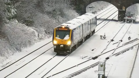 PA Media A wide shot of a yellow train travelling through snow. The track is covered with snow, and the train is covered with snow.
