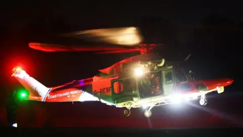 Coastguard helicopter at night with search light on and coastguard team at the door of the aircraft.