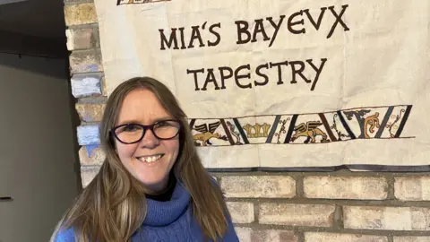 Mia Hansson has long brown hair and is wearing glasses and a blue turtleneck jumper. She is standing in front of a sign which says "Mia's Bayeux Tapestry".