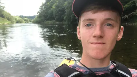 Leigh Day Thomas Garland, who has short ginger hair and freckles and is wearing a red baseball cap, a red and grey t shirt and a life jacket. He is in his canoe, which is not visible, but you can see the water and greenery of the River Wye behind him.